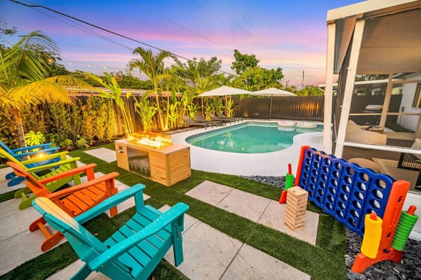 Backyard with turquoise pool, colorful Adirondack chairs, fire pit, games, tropical plants, and modern house at dusk