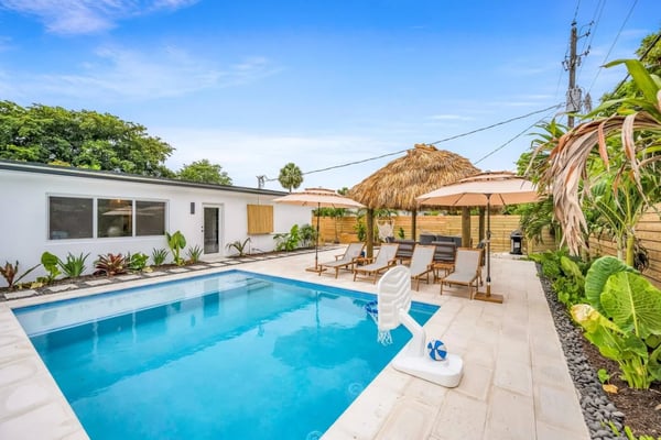 Backyard with rectangular swimming pool, lounge chairs under thatched umbrellas, white modern house, and tropical landscaping