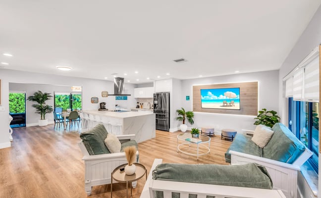 Open-plan living room with turquoise sectional sofa, dining area, TV displaying tropical beach scene, and glass doors opening to patio with plants